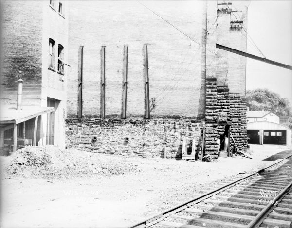 View across railroad tracks towards the back side of the Fauerbach Brewery Building, 651-653 Williamson Street. A man is standing just inside a doorway at the back of the building. A Trachte building with open doors is behind the brewery on the right.