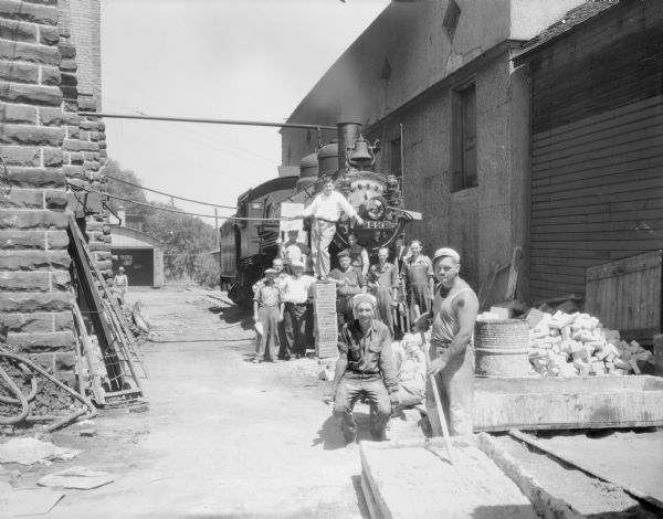 Group portrait of eleven workers installing Plibrico Jointless Firebrick in front of a steam locomotive parked beside Fauerbach Brewery and used to provide steam power while boiler was being repaired.