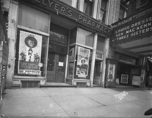 Orpheum advertisement for "The Sap from Syracuse", and a Capitol Theatre advertisement for "Let Us Be Gay" in Collyer's Pharmacy window in the Mayer Building at 14 E. Mifflin Street next to the Strand Theatre. The Strand marquee has a sign for "Three Sisters" at 16 E. Mifflin Street.