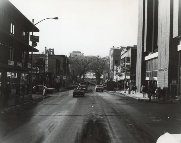 View down center of State Street towards Park Street at the bottom of Bascom Hill, with automobile traffic moving up and down the street. On the left are storefronts at the corner of Hawthorne Court, and on the next corner is the Brathaus. The building on the right is The Towers at the corner of North Francis Street. 