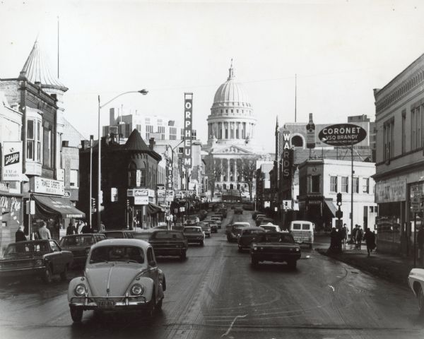 View down center of State Street, with automobile traffic moving up and down the street, towards the Wisconsin State Capitol. On the corner of W. Johnson Street on the left is the Triangle Superette. Further up the street on the left is Nicks Restaurant, and the Orpheum Theatre. Across from the Orpheum on the right side of the street is the Leath Furniture Co., the Capitol Theatre and Montgomery Ward's. 