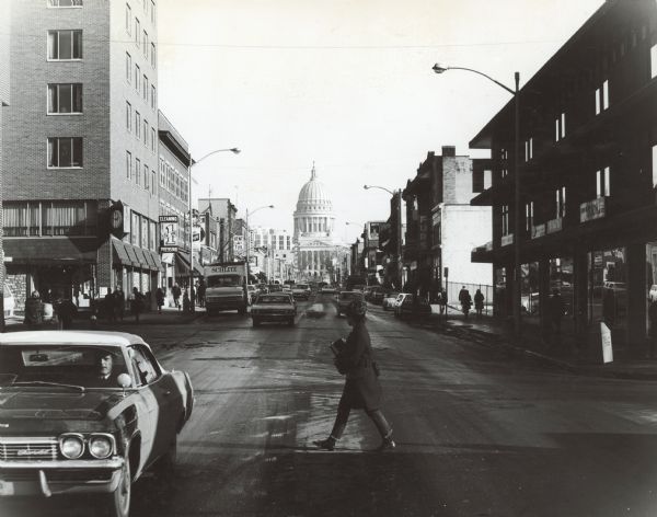 View down center of State Street towards the intersection of N. Frances Street. Further down the street on the left are signs for 1 Hour Cleaning Pressing, The Pub Tavern, and Wehrmann's Travel Shop. On the right are signs for the Yarn Bar, and Edwin O. Olson and Son Men's Clothing.