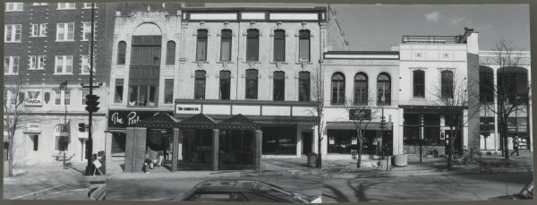 View across N. Pinckney Street looking towards towards a row of buildings, starting from the left: YWCA, The Perfume Shops (obstructed sign), The Camera Company, Panache Hair — Beauty, unknown storefront, and on the far right the Christian Science Reading Room. There is a bus stop in front of The Camera Company.