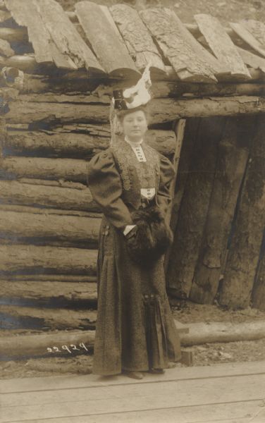 A woman is standing and posing in front of a log cabin. She is wearing a dress with floral accents and puffed sleeves, and a fascinator with a lace veil. She has her hands in a fur muff. This is a picture postcard addressed to Mr. Glenn Thompson in Madison, sent from "M.W. Mc B." Postcard message reads: "Outfitted for Easter — but don't you think the roof needs fixing? Thanks for your recent enclosure. More Later - M.W. Mc B."