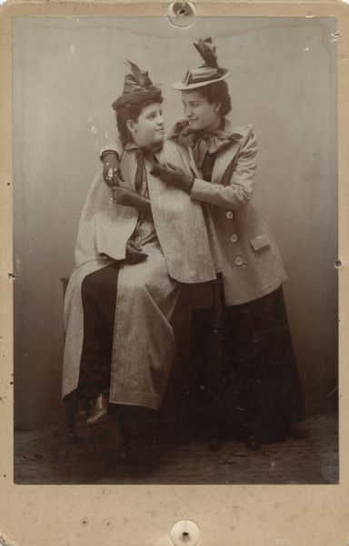 Two unidentified women posing together. The woman on the left is sitting on a table or bench, and is wearing a hat and a coat with a cape. The other woman is standing on the right and is wearing a hat and coat, and she has her arms around the other woman's shoulders.