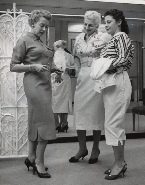Three women are standing together, with two of them wearing dresses and one woman wearing a blouse and skirt. Two of the women are looking at the third, who is looking down while holding onto the open belt of her dress. There is a mirror reflecting them in the background. Caption reads: "<b>Members of the Elm Grove Women's club</b> chose gowns which they will model at their club's benefit style show on Apr. 24. From left are Mrs. Joseph [Etta] Bader, Mrs. Gustave [Margaret] Mader and Mrs. John [Carmen] Kizivat, all of Elm Grove. The show is scheduled to begin at 7 p.m. at Brookfield high school. Mrs. H.R. Perlick, Elm Grove, is show chairman."