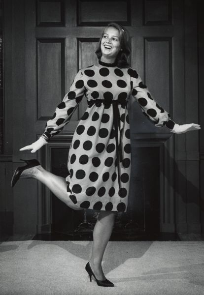 A woman is smiling while posing with her right leg lifted. She is wearing a dress and standing in front of a fireplace and wood-paneled wall. Caption reads: "BRONWYN JONES felt happy in an empire dress with king size polka dots. The sale also offers lingerie, sweaters and sports clothes for women, men and children. The price range varies."