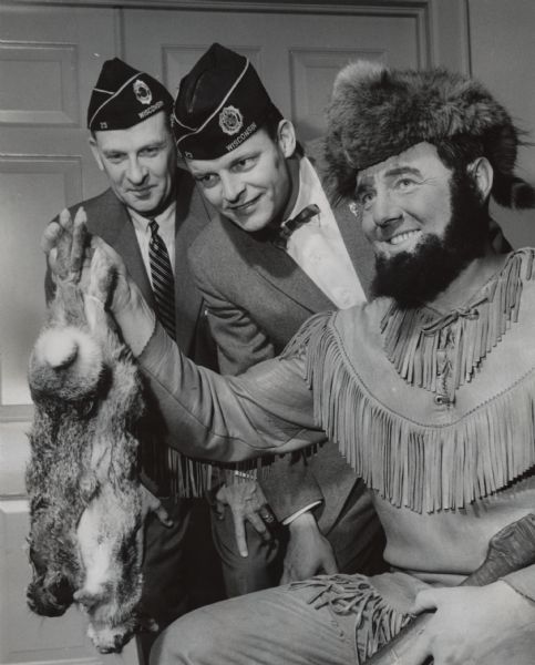 A man is posing in a leather fringed costume and fur cap, with what appears to be a fake beard. He is holding two dead animals up in his right hand. Two other men wearing American Legion caps are looking at the animals. Caption reads: <b>"A 'mighty hunter'</b> was the butt of jokes at the annual past commanders' dinner Thursday night at Cudworth American Legion Post, 1756 N. Prospect Av. Richard F. Redfield, 168 E. Willow rd., Fox Point, present commander, came in costume--as tradition demands. Past commanders Harry E. Bradley (left), 7434 Melrose Av., Wauwatosa, and Frederick F. Frick, 2851 Cleveland Park Dr., poked fun at Redfield."
