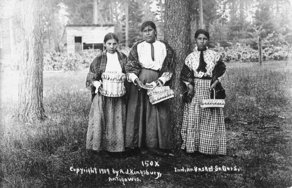 Three Potawatomi women selling baskets posing by a tree. There is a building in the background. Caption reads: "Indian Basekt Sellers."