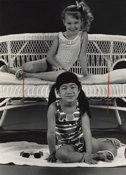 Two girls are posing while wearing swimsuits and sandals; one girl is sitting on a wicker bench, and the other girl is siting on a towel in front. Caption reads: "Swimwear (below) is off to a hot start with metallic glitter and bright colors, as in this multicolor print tank suit, about $24 from Louise Goodell, or the pale pink tank, $12 from The Children's Place. The one-piece style, or monokini, is expected to be the most popular look this summer. Neon-accented sunglasses add a fun touch."