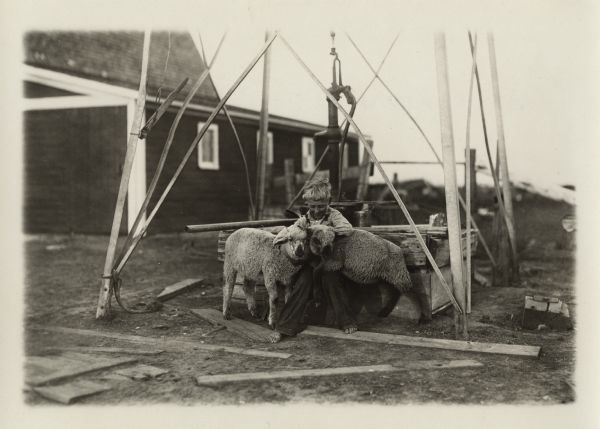 A barefoot child is holding two lambs close to him while sitting at the base of a well pump. A farm building is in the background.