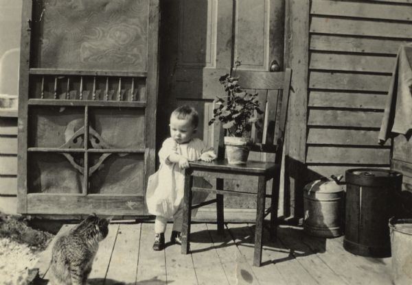 A toddler is standing outdoors on a wooden landing while holding onto a chair with a potted plant on it. A cat is sitting nearby on the left, looking back at the child. 
