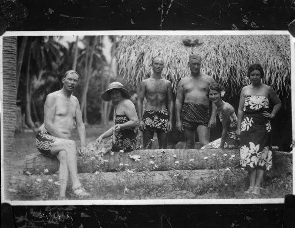 Group portrait of Professor Ross and five friends in native dress in Tahiti. Two of the people are sitting on a stack of logs, with a cat sitting between them.