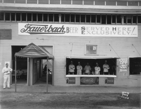 View towards the Fauerbach concession stand, with five servers, all men, standing behind the counter at the Dane County fair. There are signs for Coca-Cola and "Blatz quality soda 15 flavors."