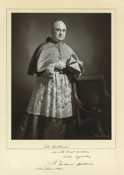 Portrait of Cardinal Francis Spellman. He is standing next to a chair and is holding a biretta (hat) in one hand and wearing his choir dress. Spellman is also wearing a large cross around his neck and a large ring on his finger. The handwritten inscription reads: "Ed Sullivan/with best wishes/and regards,/F. Cardinal Spellman/Christmas 1947."