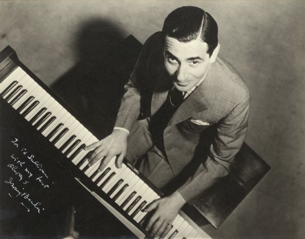 Portrait from above of Irving Berlin sitting at a piano. Berlin has his hands on the keyboard, as if playing, and is looking up at the camera. The handwritten inscription reads: "For Ed Sullivan/with my best/always/Irving Berlin."