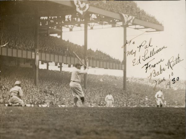 View across field towards Babe Ruth at bat during a baseball game, taken from ground level. Ruth's right side is to the camera and he is holding the bat above his head. The catcher is on his knees behind Ruth on the left, and two other men can be seen on the field. Each of the three levels of the stands are full of people and bunting is hanging from the top level. The handwritten inscription reads: "To/My Pal/Ed Sullivan/From/Babe Ruth/Jan 10/1930."