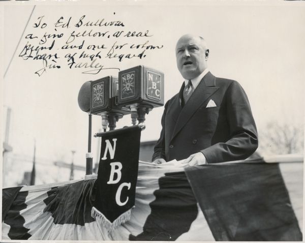 Postmaster General James Farley speaking at the ceremony to lay the cornerstone for the new post office in Haverstraw, New York. Three large microphones, including two for the NBC network, are attached to the railing in front of him. An NBC pennant is hanging from the microphone stand. The handwritten inscription reads: "To Ed Sullivan/a fine fellow, a real/friend and one for whom/I have a high regard./Jim Farley."