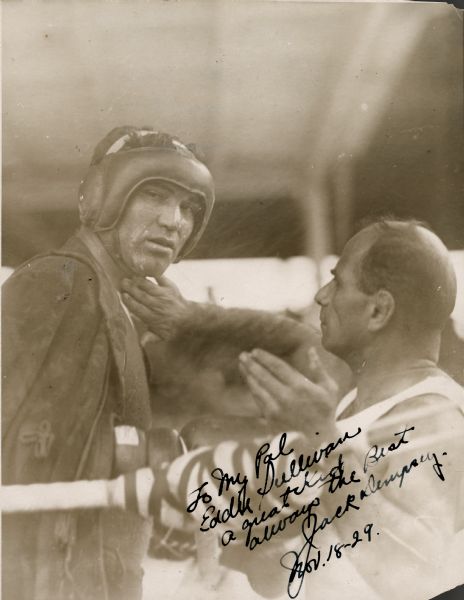 Trainer Jerry "The Greek" Luvadis is putting something on boxer Jack Dempsey's throat during a training session. Dempsey is wearing headgear and a robe and is looking toward the camera. The handwritten inscription reads: "To My Pal/Eddie Sullivan/a great kid/always the best/Jack Dempsey/Nov. 18-29."