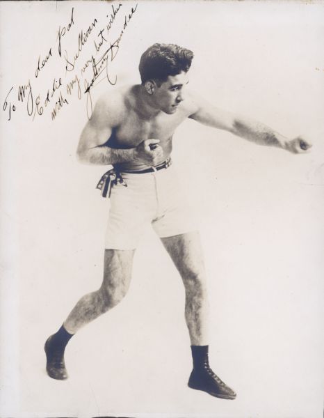 Posed portrait of boxer Johnny Dundee. He is standing with one arm extended, and the other arm bent and tucked into his chest. Both of his fists are closed. He is wearing white boxing shorts with a belt tied to one side, and black boxing shoes. The handwritten inscription reads: "To my dear pal/Eddie Sullivan/with my very best wishes/Johnny Dundee."