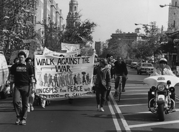 A group of people are marching up a city street holding banners, the most prominent of which reads: "Walk Against the War, Join CISPES [Committee in Solidarity with the People of El Salvador] for Peace with Justice in El Salvador." A police officer on a motorcycle is riding alongside them on the right.