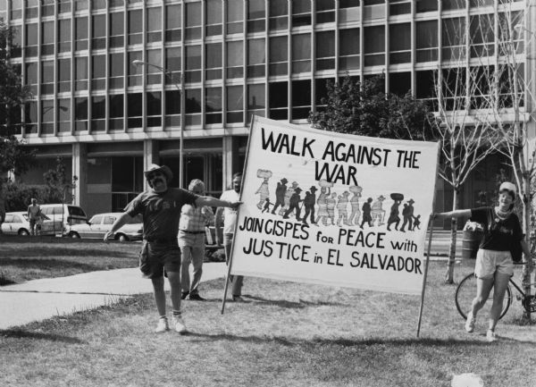 Two people are posing holding a sign that reads: "Walk Against the War, JOIN CISPES [Committee in Solidarity with the People of El Salvador] for PEACE with JUSTICE in EL SALVADOR." They are standing in front of what appears to be a large office building.