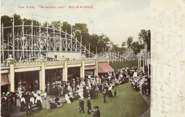 Tinted postcard showing crowds of people standing in and near shops. Behind the shops is a roller coaster. Caption reads: "The Pike, 'Wonderland' Milwaukee, Wis."