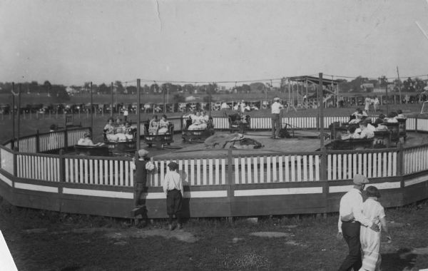 View towards groups of people riding in carts as part of an amusement park ride. Two boys in the foreground are standing and watching at the perimeter of the fence surrounding the ride. The ride operator is standing on the platform in the middle of the ride by a wheel. Automobiles are parked at the edge of the field in the background.