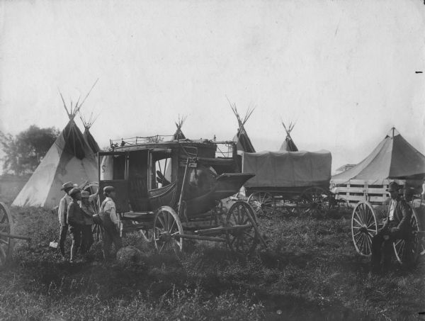 Four children on the left are standing and looking through the open door of a stagecoach, in which a man, (obscured by the side of the coach) is sitting with his legs propped up. On the far right is a man sitting on the axle of a wagon wheel. A covered wagon and several tents and tipis are in the background.