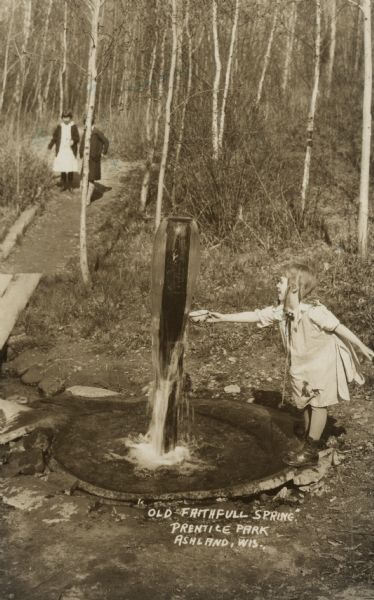 A child is holding out a bowl to a pillar from which water is cascading down into a spring. In the background, two other children are walking up a dirt path in a forest.