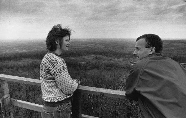 Two people are leaning against a railing overlooking at forest. Caption reads: "Rachel Imsland, 25, and Mark Evans, 28, both of Iowa City, Iowa, viewed the countryside from atop the east observation tower at Blue Mound State Park west of Madison."