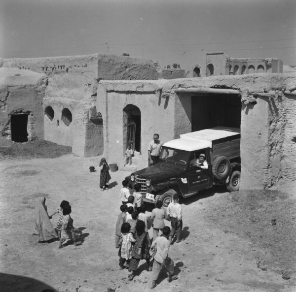 Elevated view of two men and a group of children in a courtyard. One of the men is driving a Malaria Control truck, and the other man is standing by the passenger side of the truck. A group of children are walking towards the truck, and two other children are standing near the entrance of a building.