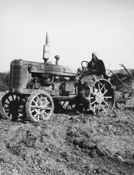 A man is driving a Super W-6 McCormick International tractor in a field. The tractor is pulling an implement, and there is a thatched-roof building in the background.