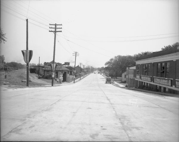 View looking east from Monroe Street, down the newly widened and paved Regent Street. Businesses include Castle and Doyle building supplies on the left (1512 Regent Street) and Wisconsin Auto Body on the right (1525 Regent Street). On the far left are railroad tracks, and the Wisconsin State Capitol is obscured by a banner attached to a power pole.