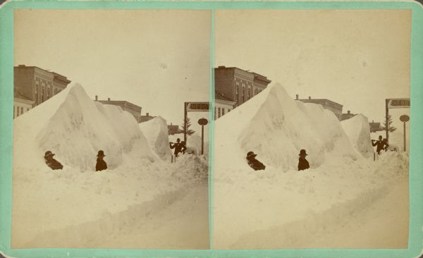 View down snowy sidewalk towards men posing in snowbanks. Another man is standing and is carrying a shovel across his shoulders. A sign over the sidewalk reads: "Sheboy[gan Tribune]." Text on back of stereograph reads: "taken from corner of 7th St." and "snow mountain 25 ft. high near Pa? House."