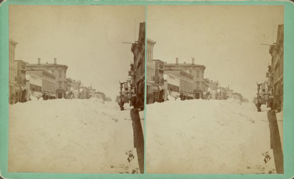 View down snow covered 8th Street. Text on back reads: "8th St. looking south, taken from Corner of Penn'a Ave." and "average height of snow 10-12 ft." Signs on some of the buildings on the left side of the street read: "Wisconsin House" and "F. Geele." On the right are many storefronts, and there is a mortar and pestle on top of a pole along the curb.