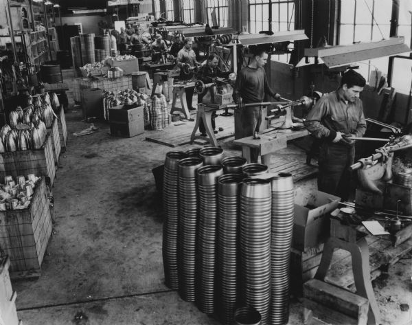 Elevated view of a machine shop, where several people are standing and working at lathes. Around the shop are piles of machined parts, all of which are cylindrical or conical.