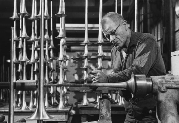 A man is working on a metal horn on a lathe. Several similar horns are stacked behind him. Caption reads: "<b>A cornet</b> slowly takes shape on a lathe at the Frank Holton & Co., Elkhorn. The firm manufactures 58 different musical horns. Howard Tess, a bell spinner for 28 years with the firm, is forming the nickel silver flare of the instrument."