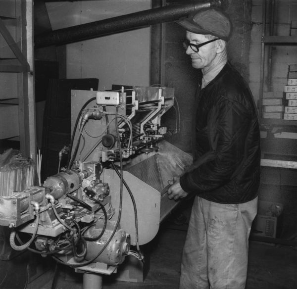 A man is standing and holding something in front of a machine. Caption reads: "ELOF ELFSTROM FEEDS NOCK MACHINE. He Designed Precision Device."