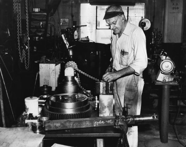 A man is standing and working in front of a machine. Caption reads: "<b>PROSPECTING DEVICE</b> - When John Yungers completes this special project, it will be possible to do prospecting work for clay beds in the Red Wing area. The Goodhue machinist, now 70, is building the device from his own plans."
