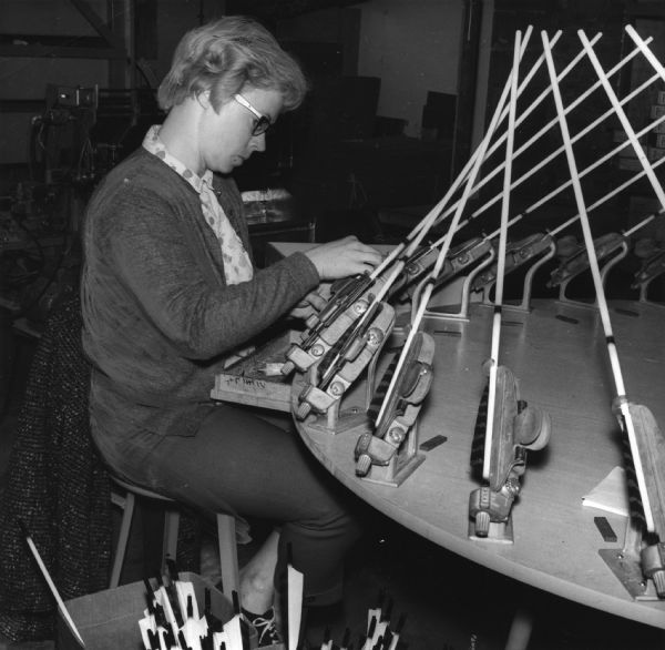 A woman is sitting at a machine where several arrow shafts are held in place. Caption reads: "ANN ELFSTROM FEATHERS THE SHAFTS. Fletching Wheel Is Exacting Operation."