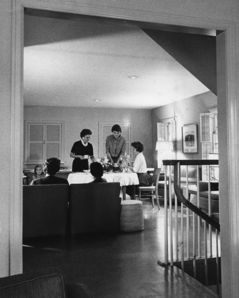 View through a doorway towrads three women sitting and standing at a table. A child and other people are sitting nearby. Caption reads: "<b>Miss Barbara Chase</b> (seated), a Kappa Alpha Theta pledge whose home is at 2620 N. Prospect Av., served coffee to Mrs. Bliss Bowman (left), Rockford, Ill., and Mrs. Walter [Kathryn] Lillyman, Beloit."