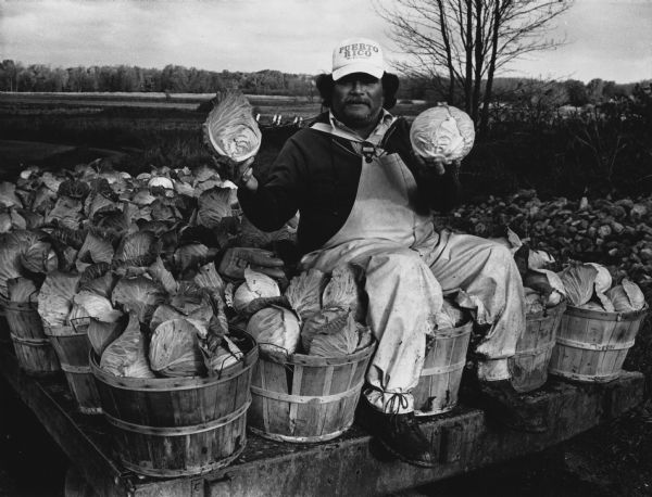 A man is sitting on a flatbed covered in baskets of cabbages, and is holding up two cabbages. He is wearing overalls, and a cap that reads: "Puerto Rico." Caption reads: "THE CABBAGE PATCH — Luis Felix, a worker at the Harold Hahm and Son Inc. farm of Mequon, proudly displayed some of the cabbages Wednesday that he helped cut at the farm. Felix is from Puerto Rico."
