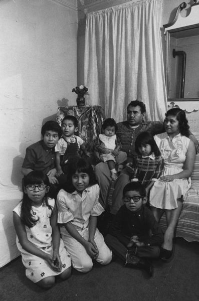 Two adults and seven children are posing together in a room. The adults are sitting on a sofa, and behind the children is a table with a vase on it. Caption reads: "There will be no Christmas tree for the Ascension Ortiz family of 709 S. 6th St. In winter all nine members must stay in the apartment's living room. They are, from left, front row: Mary Ann, 9, Alma, 10, and Paul, 6; back row: Carlo, 11, Ascension Jr., 3, Thomas, 1, and Mr. and Mrs. Ortiz with Amalia, 4."