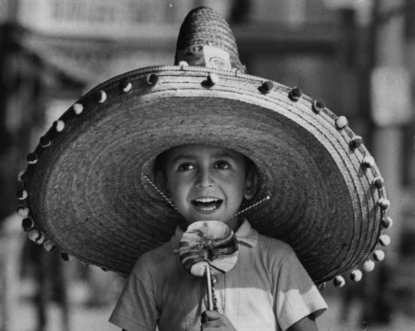 A boy is smiling while wearing a sombrero. He is holding a large lollipop in front of his mouth. Caption reads: "Wearing a big sombrero, Anthony Berrones, 4, of 818 S. 24th St., watched a Mexican independence celebration parade Sunday. The procession wound along south side streets and Wisconsin Av. on its way to the Auditorium where a dance was held. The anniversary celebration commemorated the revolt against Spain."