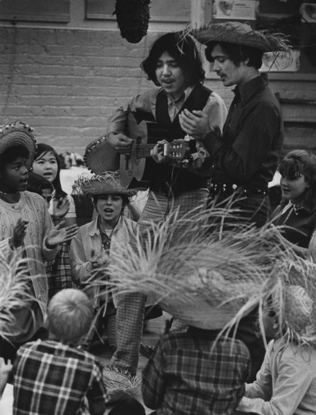 Two young men are standing among a group of children. Many are wearing sombreros. One of the adults is playing a guitar. Caption reads: "<b>FROM BELOW THE BORDER</b> - Richard Carbajam (left), 18, and Hector Altoro, 18, led a group of sixth graders in Mexican songs at the Wisconsin Ave. School, 2708 W. Wisconsin Ave. The youngsters have been studying Spanish and held a minifiesta. Carbajam, of Phoenix, Ariz., and Altoro, of Chicago, attend the University of Wisconsin-Milwaukee. They live at Carl Sandburg Hall, 3400 N. Maryland Ave."