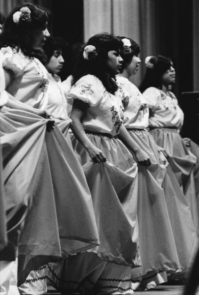 Several people in dresses are standing and holding up their skirts while  dancing. Caption reads: "A dance group, the Los Bailarimos Folkloricos de Waukesha, performed Sunday at a church festival at the Waukesha County Exposition Center."