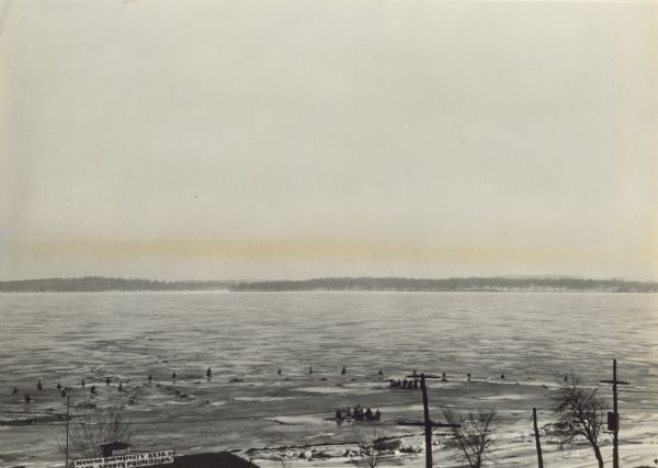 View of frozen Lake Monona from the fifth floor window of the Cardinal Hotel. Shows the "Monona Community Assn. Inc./Lake Sports Promotion!" building, including its sign on the roof, on the lake shore at the end of S. Hancock Street. There are pine trees that have been placed upright in the ice near the shoreline.