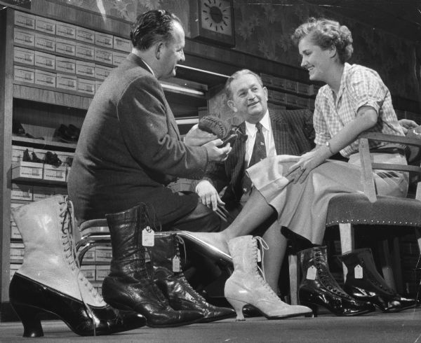 A man is holding a shoe for a woman to look at. She is resting her left foot on a shoe fitting bench. Another man is sitting nearby looking at her. They are in a shoe store, with several shoes on display in the foreground. Caption reads: "<b>Popular footwear of women</b> through the years is on display at a shoe store at 2701 W. Vliet St., in connection with the 100th anniversary celebration of Vliet St. George M. Deig (left), president of the Vliet Street Advancement association who got the exhibit together, tried a pair of the old shoes on Miss Kay Lange, 17, of 816 N. 76th St., Wauwatosa, Tuesday. Joseph Branovan, who operates the store, looked on. "