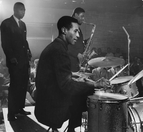 Three musicians are on a stage; an audience is in the background. One man in the foreground is playing the drums, another man is playing a tenor saxophone, and one man is holding a trumpet. Caption reads: "Progressive jazz came to the University of Wisconsin-Milwaukee campus Monday night. The Max Roach quintet played before some 600 persons who jammed the lounge of the new union at 2200 E. Kenwood Blvd. Featured in the combo were Kenny Dorham, trumpet; Sonny Rollins, saxophone; George Morrow, bass; Billy Wallace, piano, and Roach, one of jazzdom's greatest, on drums. The new UWM chapter of the National Jazz fraternity sponsored the concert."